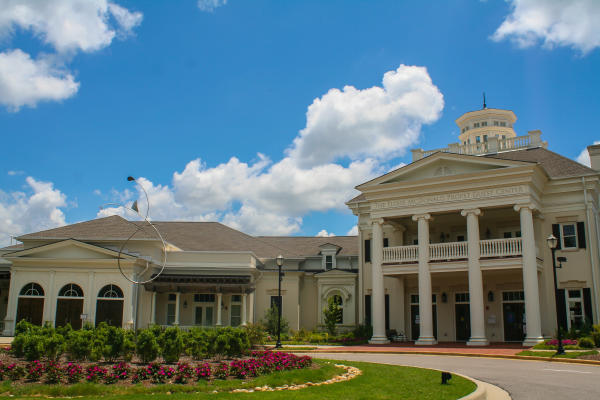 The Eloise McDonald Propst Guest Center at Huntsville Botanical Garden stands beneath a bright blue sky with large white clouds. The elegant building features tall columns, landscaped flower beds, and a welcoming entrance surrounded by lush gardens.