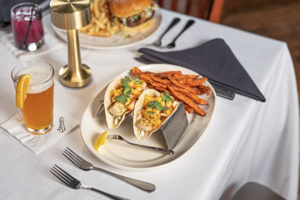 Fish tacos topped with mango salsa and cilantro are served with sweet potato fries on a white tablecloth at Connors Steak & Seafood. A cocktail garnished with an orange slice sits beside the meal, while a burger and fries are visible in the background.