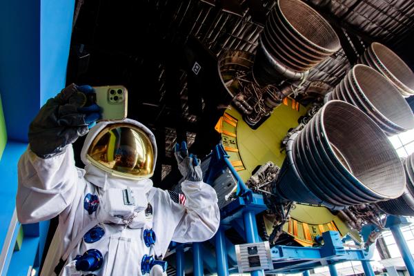 A visitor wearing a NASA astronaut suit takes a selfie beneath the massive Saturn V rocket engines inside the U.S. Space & Rocket Center in Huntsville, with the towering rocket display filling the background of the museum exhibit.
