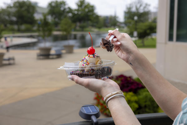A person holds a brownie sundae topped with vanilla ice cream, rainbow sprinkles, and a cherry from The Cozy Cow. A fork lifts a bite of brownie while Big Spring Park and its pond are softly blurred in the background.
