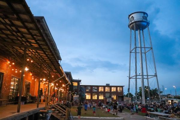 A crowd gathers outside Lowe Mill ARTS & Entertainment in Huntsville during an evening outdoor concert, with string lights glowing along the historic brick building and the iconic water tower rising above the lawn as families and visitors enjoy live music at sunset.