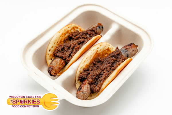 Two sausage tacos served in a compostable takeout container, each featuring a grilled bratwurst wrapped in a folded mini pancake and topped with a generous layer of chocolate chili. A “Sporkies” Wisconsin State Fair Food Competition logo appears in the bottom left corner.
