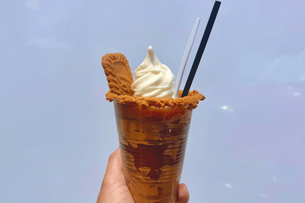 A hand holds a caramel-drizzled beverage topped with soft-serve ice cream, a cookie and cookie crumbs against a blue sky.