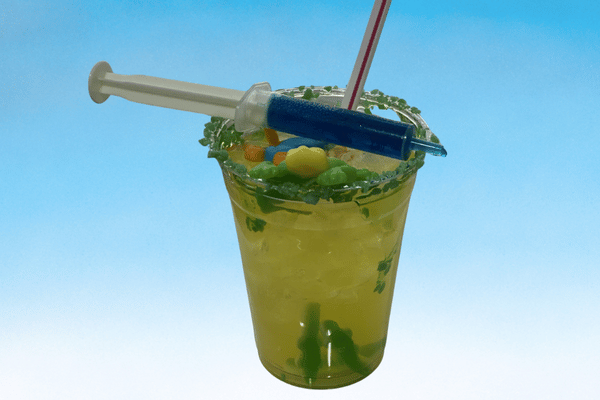 A bright green iced drink garnished with gummy candies, a sugar-coated rim and a syringe filled with blue liquid.