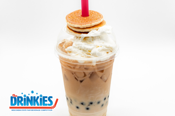 A clear plastic cup filled with iced coffee or milk tea topped with whipped cream and tapioca pearls at the bottom. A bright red straw pokes through the lid, stacked with three mini pancakes. The background is a gradient vignette with spotlight-style lighting on the drink.