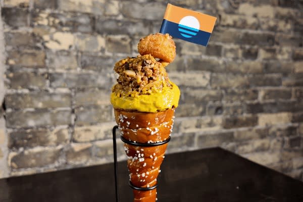 A soft pretzel cone filled with bright yellow cheese sauce and topped with chopped meat, a fried cheese curd and a small flag.