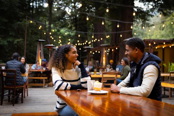 This is an image of a couple eating outdoors at Fernwood Campground & Resort in Big Sur. The couple are sitting across from each other on a glossy wooden table. The outdoor seating is beneath lush green redwood trees with yellow string lights hanging above them. The couple are looking at each other and smiling with two drinks in front of them.