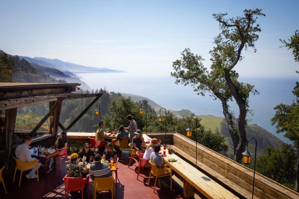 This is an image of the patio at Nepenthe in Big Sur overlooking the coastal cliffs and ocean. Various groups of people sit at tables while they eat and lookout at the views.