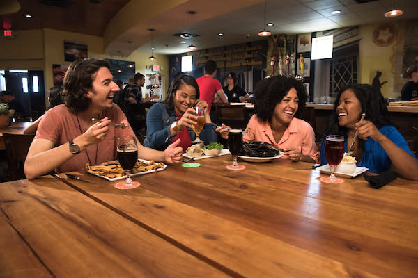A group of four friends sits at a wooden restaurant table, laughing and enjoying meals at Brewery Ommegang.