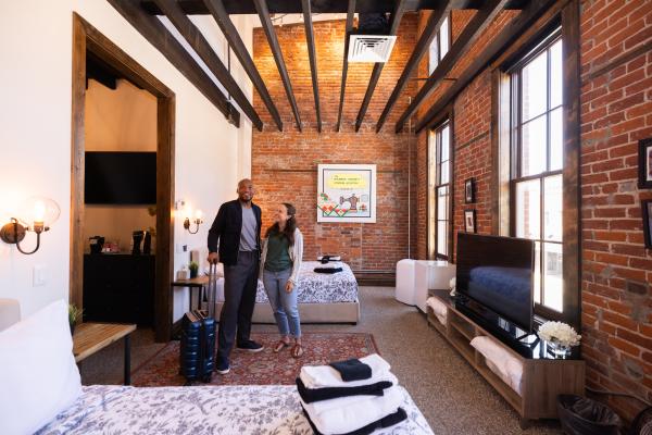 A man and a woman stand in a hotel room at the 1857. Art is hung on the walls, the beds are made, and the window have no curtains, letting natural light in.