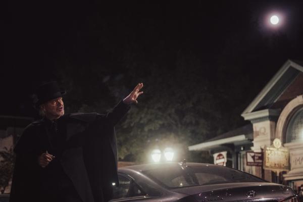 In the dark of the night, a man in all black with a top hat and cape gestures to the old Market House in downtown Paducah, which is reflected under the light of a full moon.