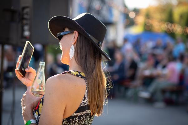 A woman in a cowboy hat and Halloween earrings holds a drink and takes pictures of live music with her phone outside at Maiden Alley Cinema's Oktoberfest.