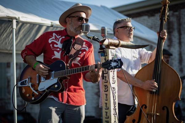 Two men play music outdoors at Oktoberfest in front of a large tent and a microphone. One is wearing a button-down white shirt and playing an upright bass and the other is wearing a cowboy hat and red Western shirt and playing an electric guitar.
