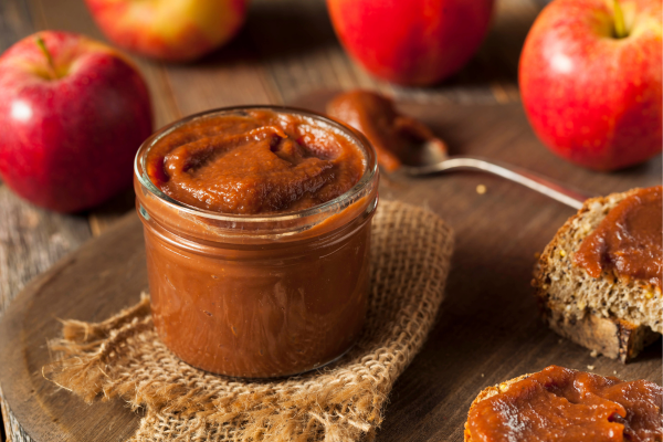 Jar of apple butter on wooden surface with apples and spoon in the background.