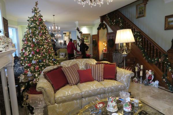 Home decorated for Christmas with a Christmas tree on the left and sofa in the middle of the room with a table with snow globes on it. The staircase has garland wrapped around and there are some people chatting in the back room