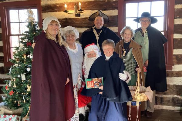 A small group of women and men stand inside a log cabin. They aredressed in 18th-century clothes and surround a traditionally dressed Santa at a holiday celebration at the Daniel Boone Homestead.