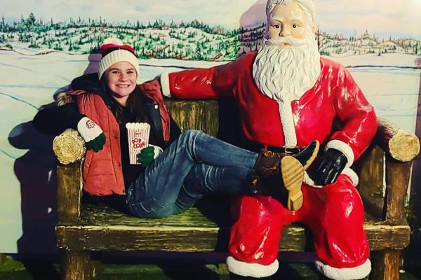 A teenage girl clad in cold-weather gear relaxes on a bench next to a sitting Santa. She is resting her crossed feet on Santa's leg while holding a box of popcorn. Stenciled above the duo are the words, Koziar's Christmas Village.