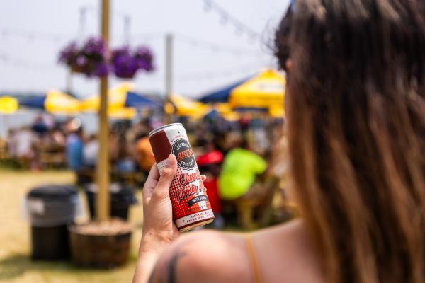 Woman holding a canned Bloody Mary while at the Lake Andrea Beer Garden. Picnic tables and flowers are seen in the background.