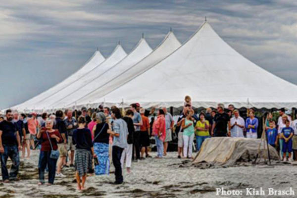 People gathered around white tents on a beach for a clambake