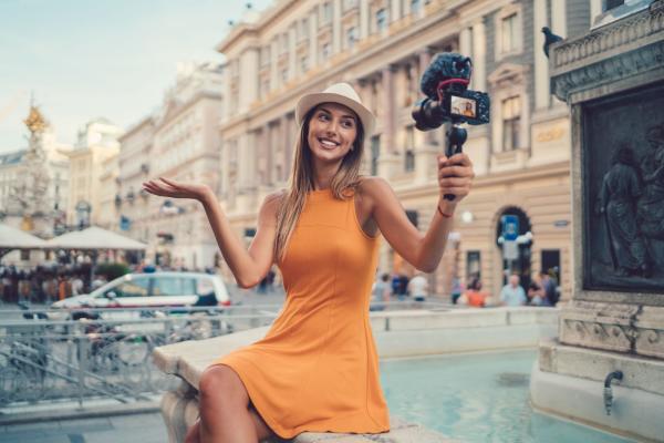 A woman in an orange sundress and hat sits on the lip of a fountain taking a video of herself