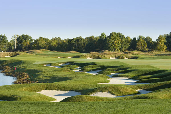 A landscaped golf course with rolling green fairways, white sand bunkers, a water feature, and trees in the background.