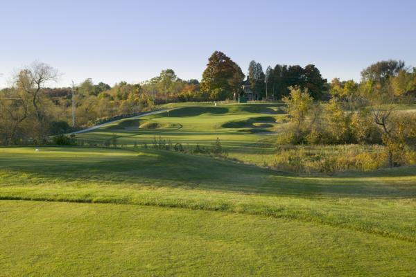 A golf course fairway leading toward a green with sand bunkers, surrounded by trees and fall foliage.