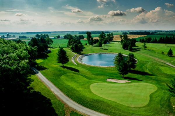 An aerial view of a golf course with a green, sand bunker, cart path, trees, a pond, and farmland in the distance.