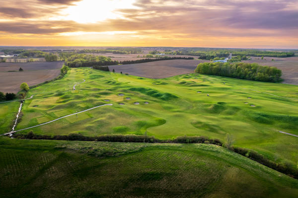 An aerial view of a rolling golf course with green fairways, sand bunkers, cart paths, and farmland in the distance at sunset.