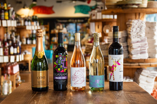 A selection of vibrant wine bottles, including sparkling and varietal wines from Mexico, beautifully displayed on a wooden table at Alta Baja Market in Santa Ana, with shelves of artisanal goods and decor in the background.
