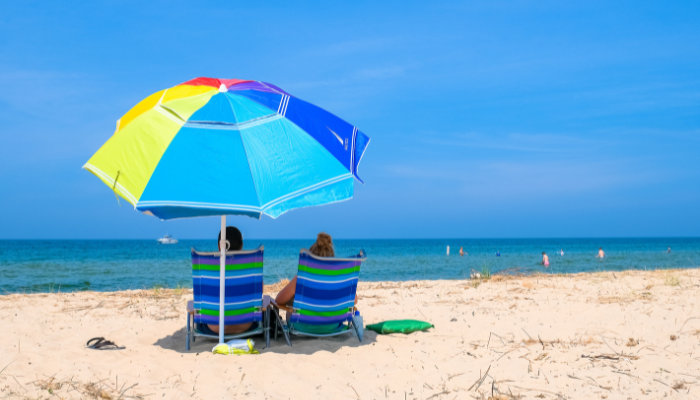 Couple sits in striped beach chairs, beneath brightly colored Beach umbrella looking toward the blue waters of lake michigan at muskegon state park beach