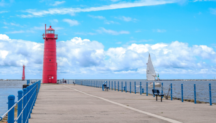 Two red light towers stand against blue sky and white clouds near the entrance to the muskegon channel. to the right of one of  the piers is  a sailboat