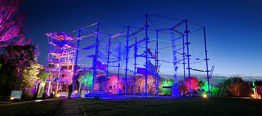 Large illuminated high ropes structure glowing in blue, purple and pink lights at dusk, with colourful lighting across surrounding trees and parkland at 270 Climbing in the Cotswolds.