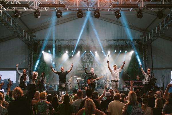Band performing on stage at the Dublin Irish Festival, with musicians holding instruments and engaging the crowd under colorful stage lights in a large tented venue.