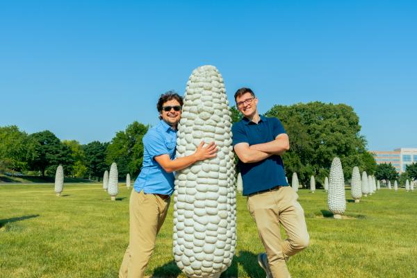 Two men smiling and posing with a large corn-shaped sculpture in a field surrounded by similar corn statues. The background features a clear blue sky, trees, and grassy open space.