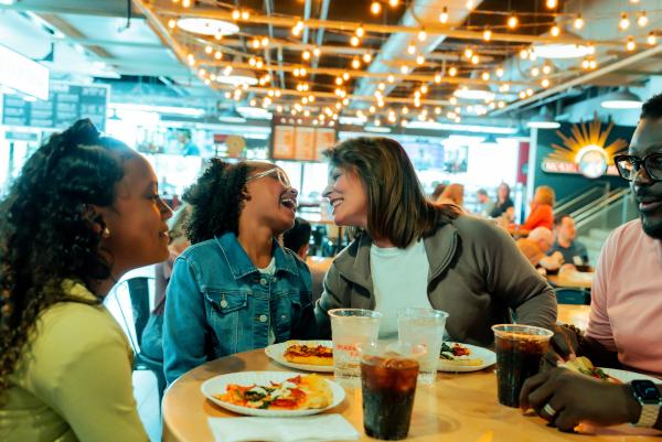 A family shares laughter and pizza at a lively indoor food hall with string lights and a vibrant atmosphere in the background.