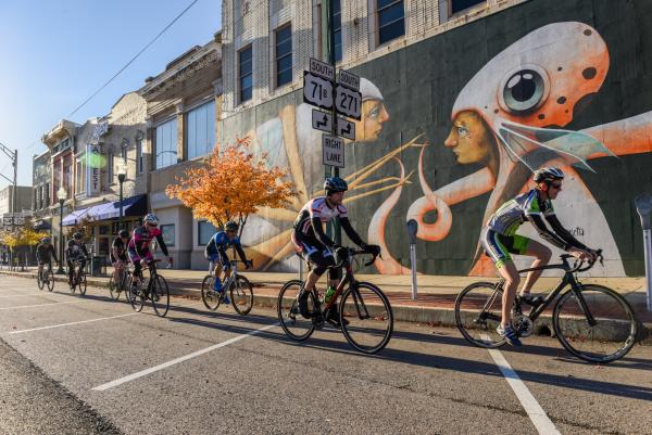Road bikers bike past a colorful mural painted on a brick building in Fort Smith.