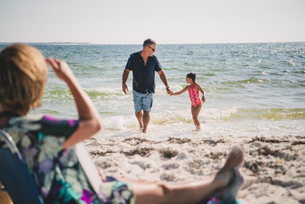 father and daughter walk hand in hand out of the water as mom watches from up on the beach.