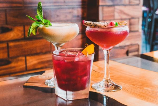 Three cocktails sit on a wooden serving plate in front of a brick wall at Brick & Bourboun in Maple Grove