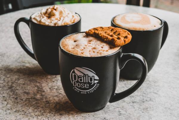 Three coffee mugs filled with foamy drinks sit on a circular table in a triangular shape. The mug in front says "Daily Dose Cafe and Espresso"