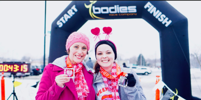 Two women decked out in Valentine's Day attire smile with medals at the finish line of a race
