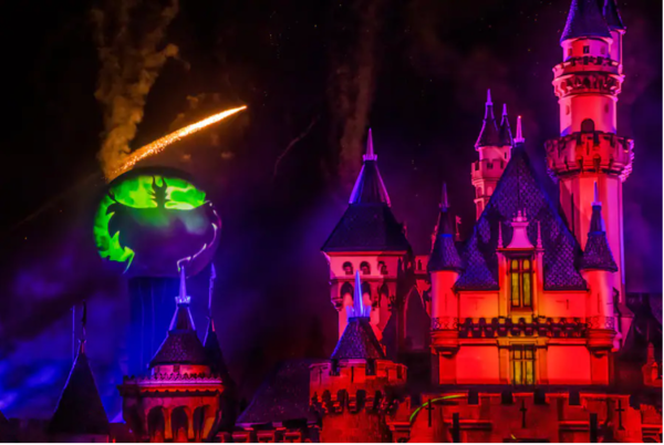 Image of the Halloween Screams Fireworks show. Sleeping Beauty's Castle can be seen in the image, lit up in a pink/red/orange hue. Fireworks and projections can be seen to the left of the castle.