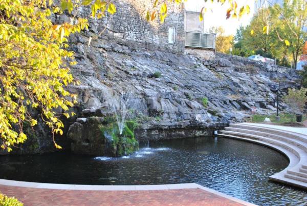 Big Spring Park’s historic natural spring flows beneath a rocky limestone bluff in downtown Huntsville, with calm water, stone seating steps, and trees surrounding one of the city’s most iconic gathering spaces.
