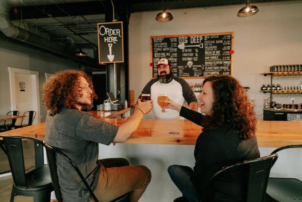 Couple cheers holding craft beers at the bar with the brewery behind the bar.