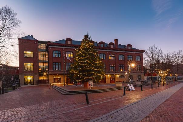 A large Christmas tree is covered in white lights in front of a historic brick depot building at dusk.