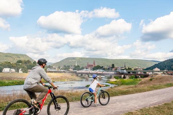 Adult and child riding bicycles along the C&O Canal Towpath in Cumberland, Maryland, with a river, bridge, downtown buildings, and green mountains and Western Maryland Scenic Railroad steam engine in the background under a blue sky.
