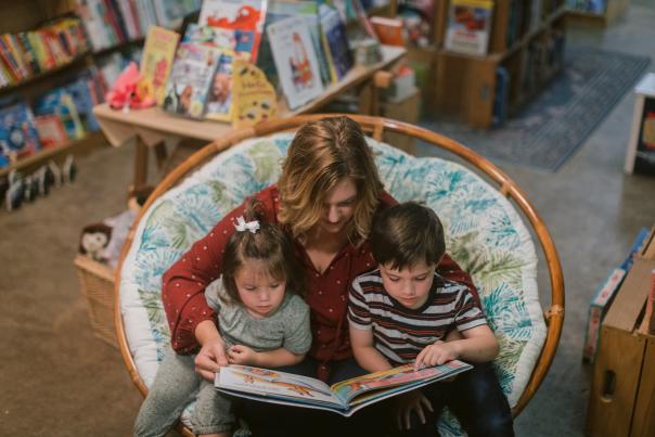 A mom in a red shirt sits in a basket chair, with an open book and her two children on her lap.