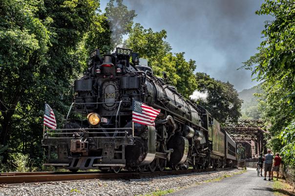 A large black steam engine runs along train track in the summer with smoke billowing from front and two American flags on either side of the engine.