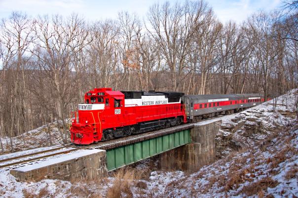 Red Western Maryland Scenic Railroad train crossing a bridge through a snowy forest landscape.
