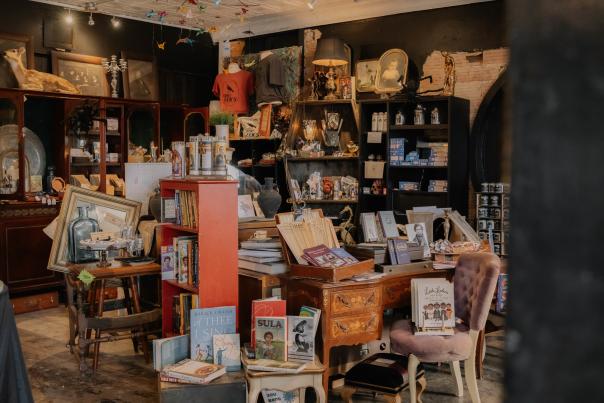 A vintage and eclectic bookstore with antique furniture, unique decor, and an array of books and collectibles. The space features a mix of wooden shelves filled with literature, a red bookcase with curated selections, and a beautifully carved wooden desk displaying books, pamphlets, and trinkets. Ornate chairs, framed artwork, and historical memorabilia contribute to the shop’s nostalgic ambiance. The dark walls, exposed brick, and warm lighting create an inviting atmosphere, while suspended colorful paper cranes add a whimsical touch.