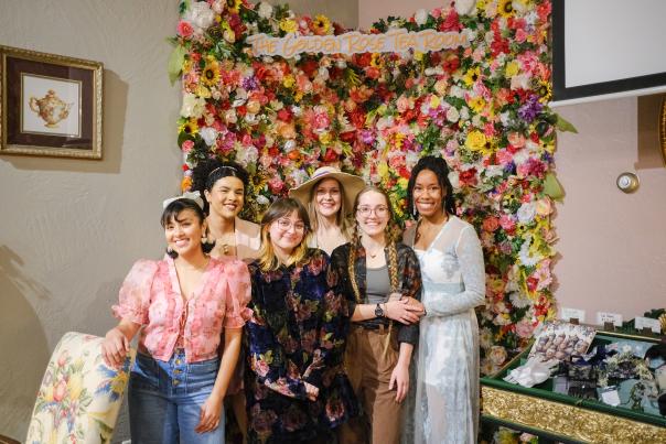 A group of six women dressed in elegant and vintage-inspired outfits pose together in front of a floral wall backdrop with a sign reading "The Golden Rose Tea Room." They are smiling and standing close together in a warmly lit tea room setting. The background features a framed picture of a teapot, a floral-upholstered chair, and a display table with accessories and trinkets.